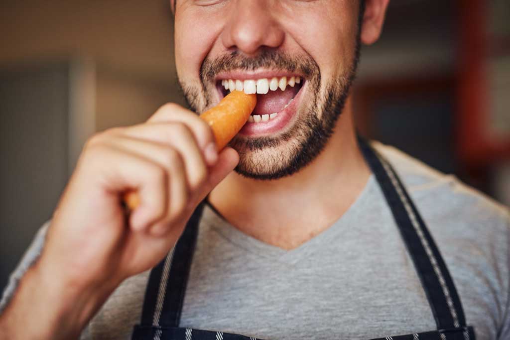 Young-man-eating-a-carrot-in-his-kitchen-at-home-to-help-prevent-tonsil-stones