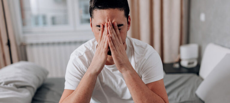 man-sitting-on-the-edge-of-a-bed-rubbing-his-face